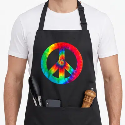 Peace sign tie-dye apron - Large peace symbol filled with red orange yellow green blue purple tie-dye pattern — shown on a black cotton canvas apron against a white background