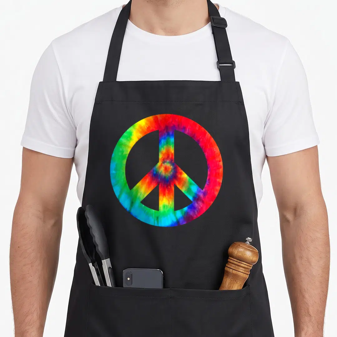 Peace sign tie-dye apron - Large peace symbol filled with red orange yellow green blue purple tie-dye pattern — shown on a black cotton canvas apron against a white background