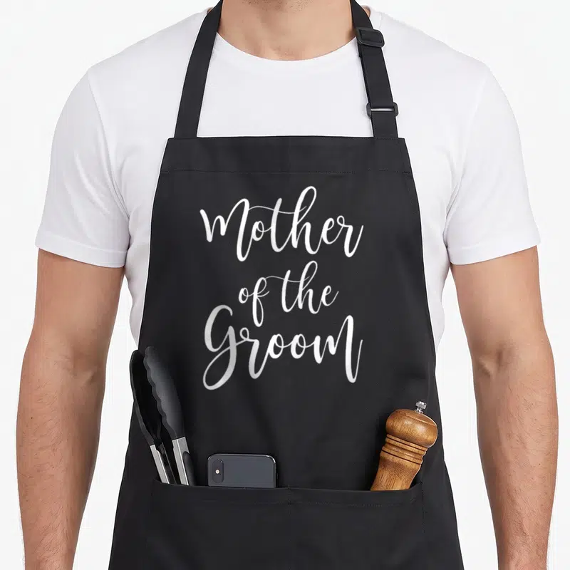 Mother groom bridal apron - White cursive text Mother of the Groom stacked on black fabric — shown on a black cotton canvas apron against a white background