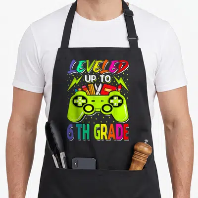 Level love grade apron - Lime green game controller with school supplies and multicolored LEVELED UP TO 6TH GRADE text — shown on a black cotton canvas apron against a white background