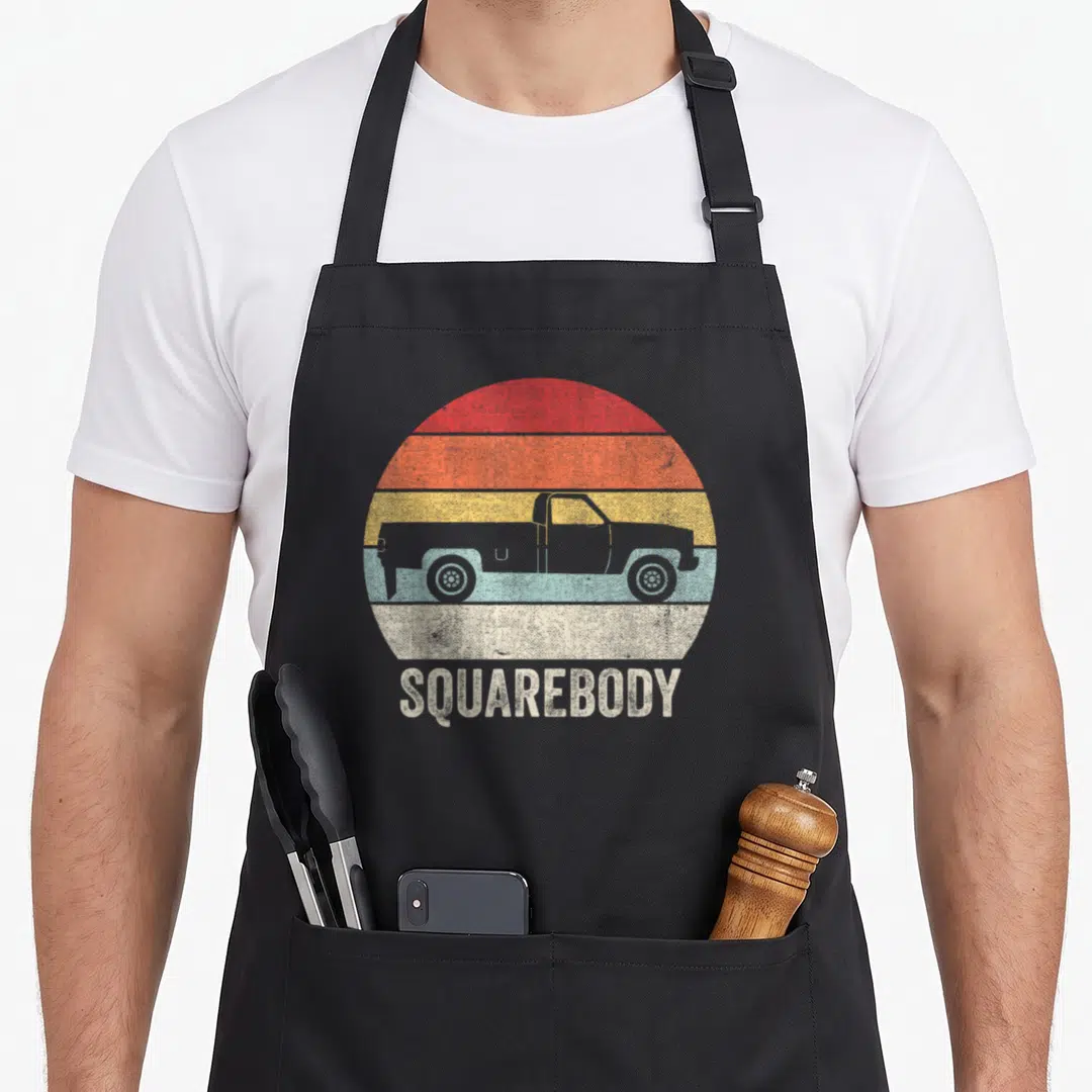 Squarebody pickup truck apron - Grey truck silhouette on red orange yellow blue striped circle with text — shown on a black cotton canvas apron against a white background