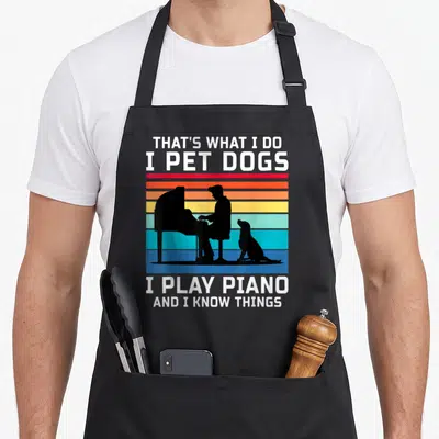 Pet piano apron - Text reading I Play Piano next to black silhouette of man and dog on colored stripes — shown on a black cotton canvas apron against a white background