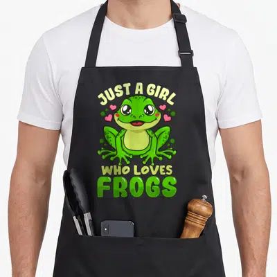 Funny frog aprons - Cartoon green frog with yellow text Just a Girl Who Loves Frogs and pink hearts — shown on a black cotton canvas apron against a white background