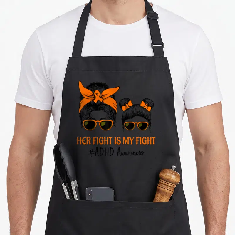 Fight messy aprons - Line art of two female heads with orange sunglasses and ribbons above ADHD Awareness text — shown on a black cotton canvas apron against a white background