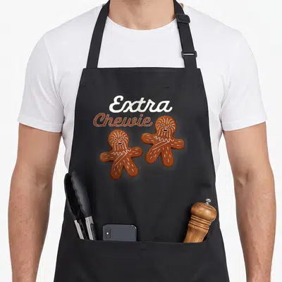 Extra gingerbread apron - Two brown gingerbread men with white icing fur details and Extra Chewie text — shown on a black cotton canvas apron against a white background