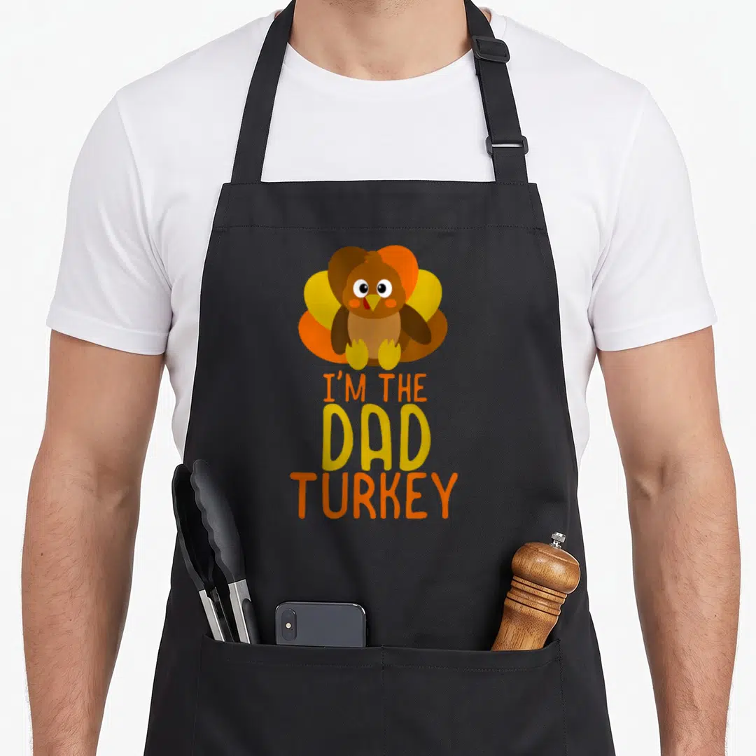 Funny family thanksgiving apron - Cartoon turkey above stacked I'M THE DAD TURKEY text in brown orange yellow — shown on a black cotton canvas apron against a white background