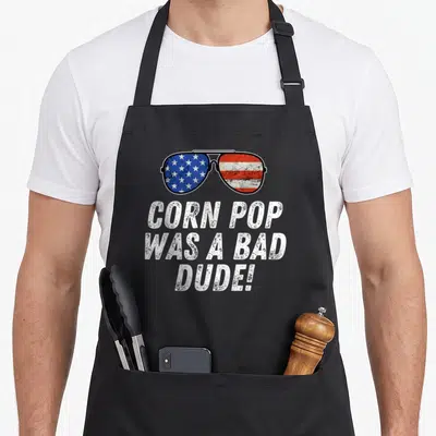 Joe dude apron - Sunglasses with flag lenses and text reading CORN POP WAS A BAD DUDE! — shown on a black cotton canvas apron against a white background
