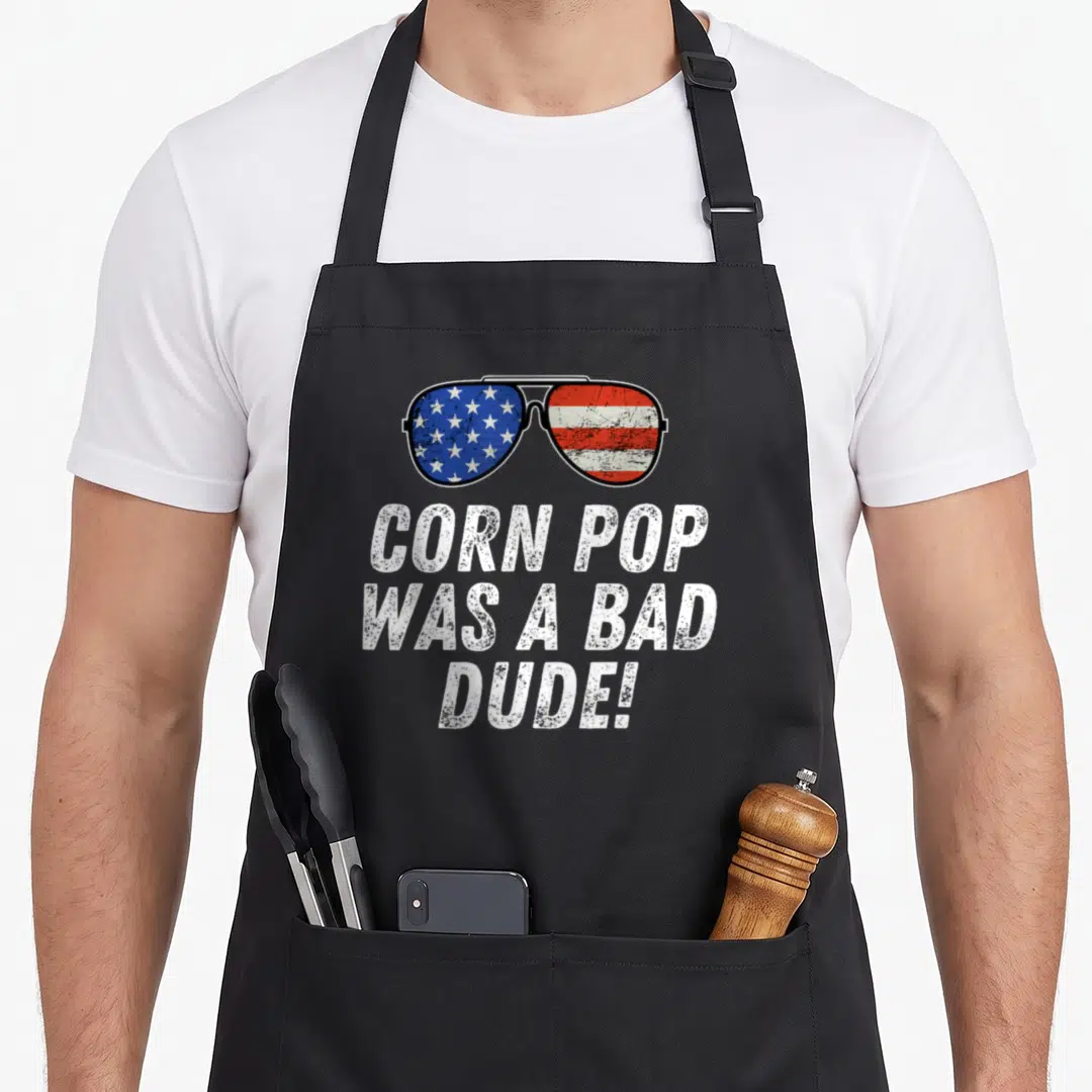 Joe dude apron - Sunglasses with flag lenses and text reading CORN POP WAS A BAD DUDE! — shown on a black cotton canvas apron against a white background
