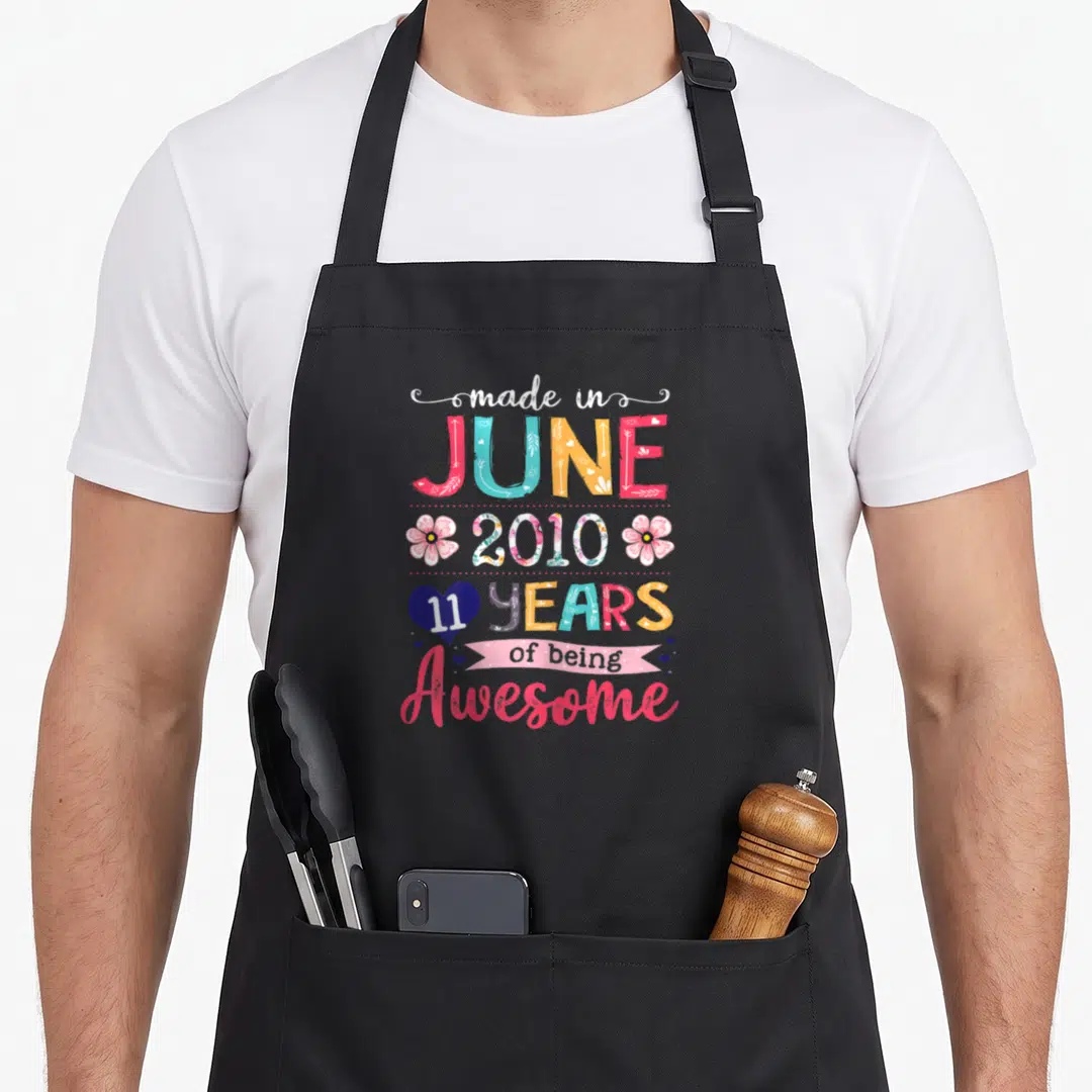 June 11th birthday apron - Stacked colorful text reading made in JUNE 2010 11 YEARS of being Awesome with flowers — shown on a black cotton canvas apron against a white background