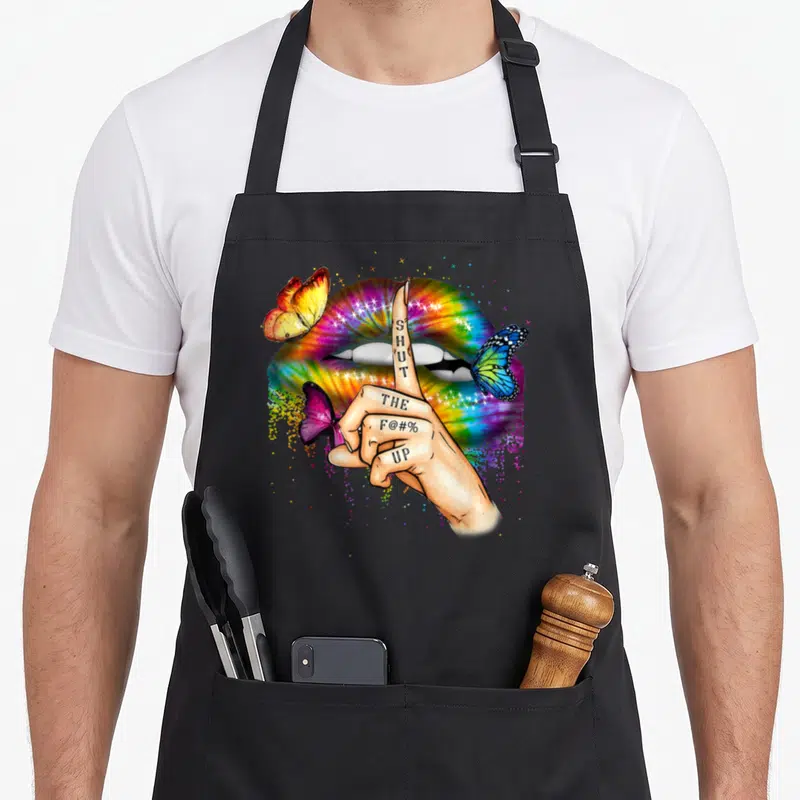 Trippy tie-dye apron - Hand with SHUT THE F@#% UP tattoo in front of rainbow lips and three butterflies — shown on a black cotton canvas apron against a white background