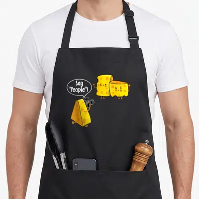 Cheese milk aprons - Three cartoon cheese wedges, one holding camera, saying Say People in speech bubble — shown on a black cotton canvas apron against a white background