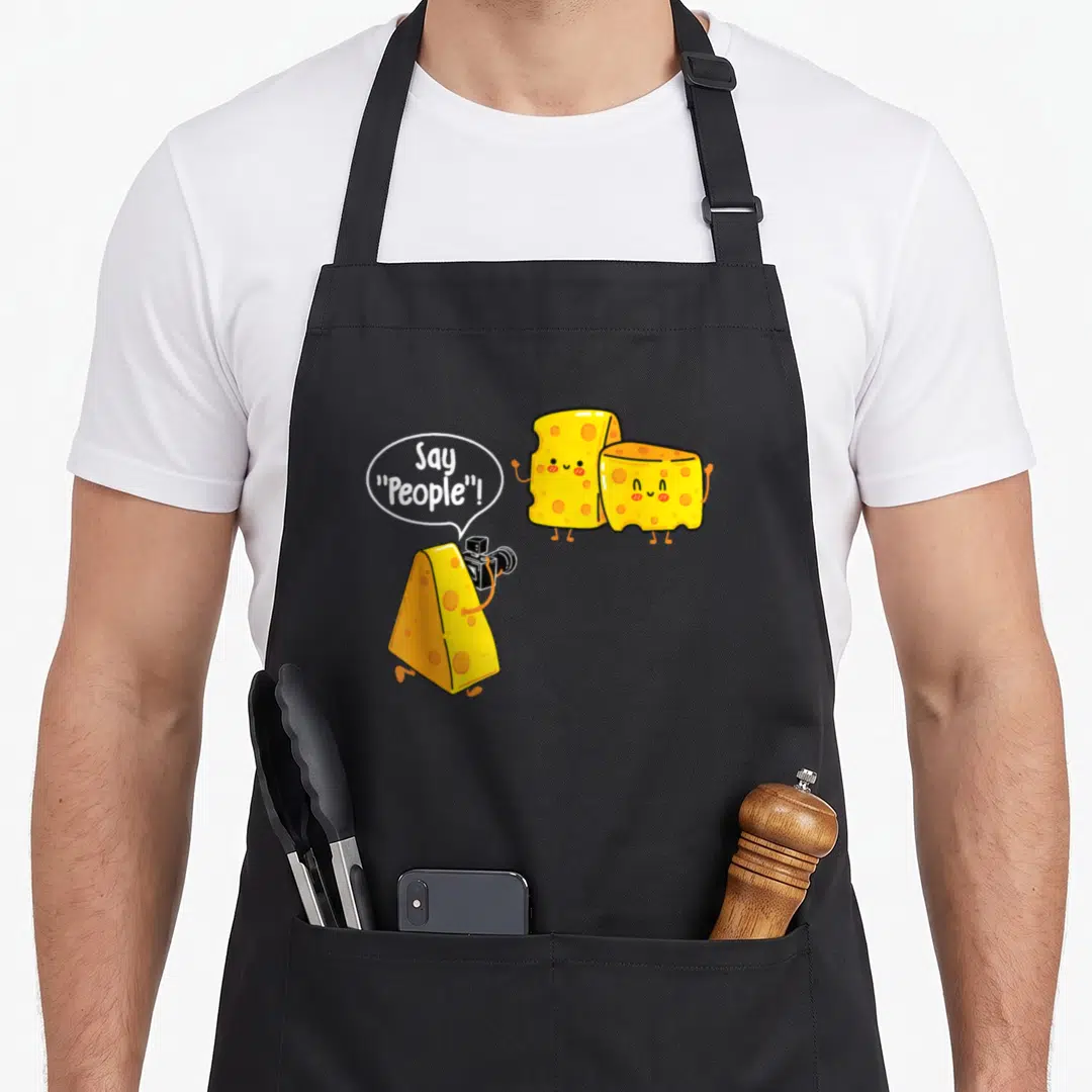 Cheese milk aprons - Three cartoon cheese wedges, one holding camera, saying Say People in speech bubble — shown on a black cotton canvas apron against a white background