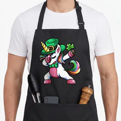 Dabbing unicorn st patrick day aprons - Cartoon white unicorn in green hat and pink clothes dabbing while holding — shown on a black cotton canvas apron against a white background