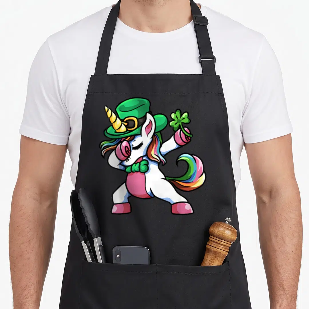 Dabbing unicorn st patrick day aprons - Cartoon white unicorn in green hat and pink clothes dabbing while holding — shown on a black cotton canvas apron against a white background