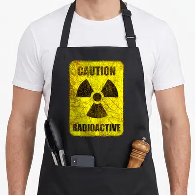 Hazard radiation aprons - Distressed yellow sign with black CAUTION RADIOACTIVE text and trefoil symbol — shown on a black cotton canvas apron against a white background