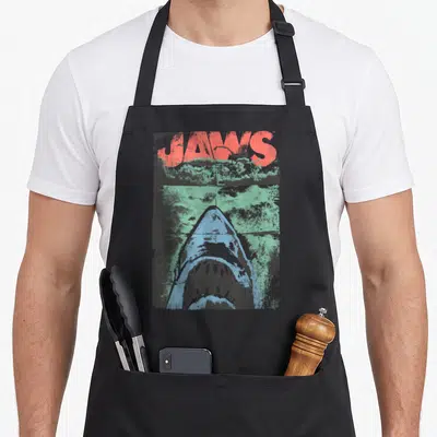 Movie distressed apron - JAWS text in red above a blue shark head in green water with distressed texture — shown on a black cotton canvas apron against a white background