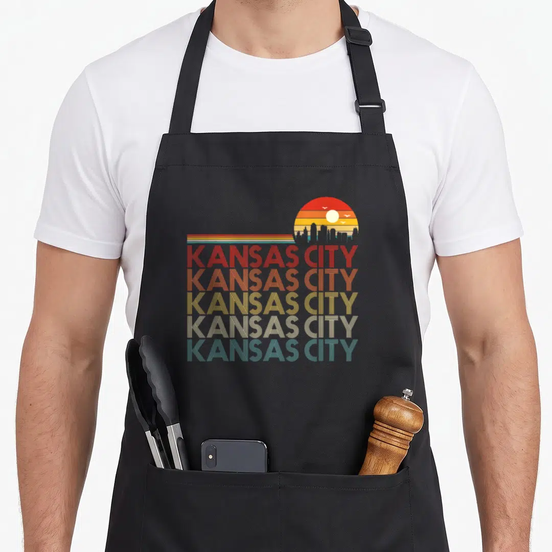 Missouri 80s apron - Five stacked KANSAS CITY text lines in red orange yellow white and blue-grey below a retro sunset — shown on a black cotton canvas apron against a white background