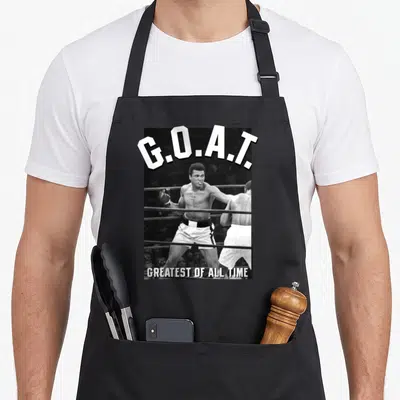 Muhammad greatest apron - Arched G.O.A.T. text above black white photo of two boxers with Greatest of All Time below — shown on a black cotton canvas apron against a white background