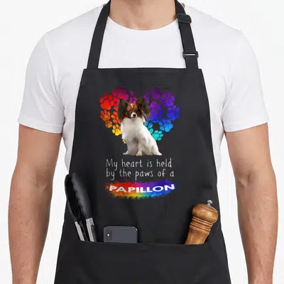 Held papillon apron - Sitting Papillon dog in front of rainbow paw print heart with stacked text below — shown on a black cotton canvas apron against a white background