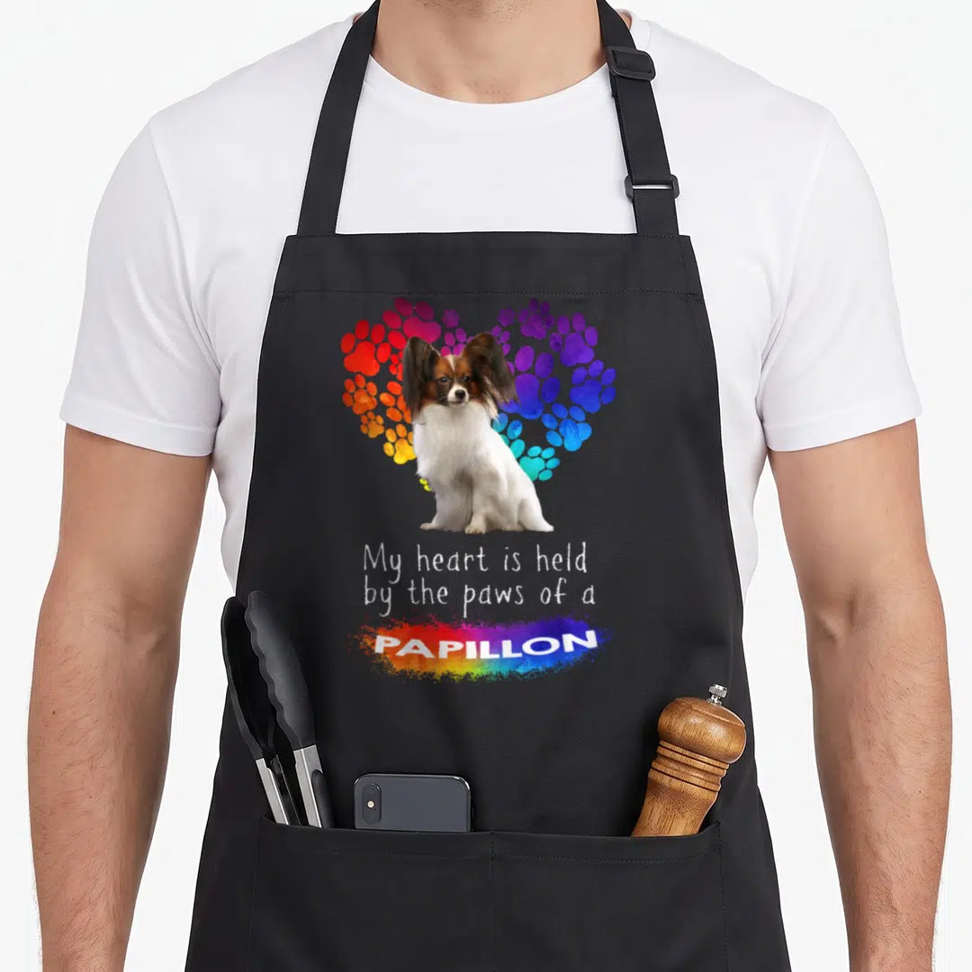 Held papillon apron - Sitting Papillon dog in front of rainbow paw print heart with stacked text below — shown on a black cotton canvas apron against a white background