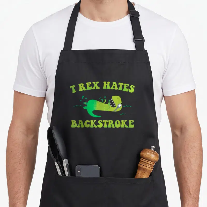 Hate dinosaur apron - Green cartoon T-Rex swimming on back with goggles and text T REX HATES BACKSTROKE — shown on a black cotton canvas apron against a white background