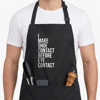 Contact shoe apron - White stacked text reading I MAKE SHOE CONTACT BEFORE EYE CONTACT on grey background — shown on a black cotton canvas apron against a white background