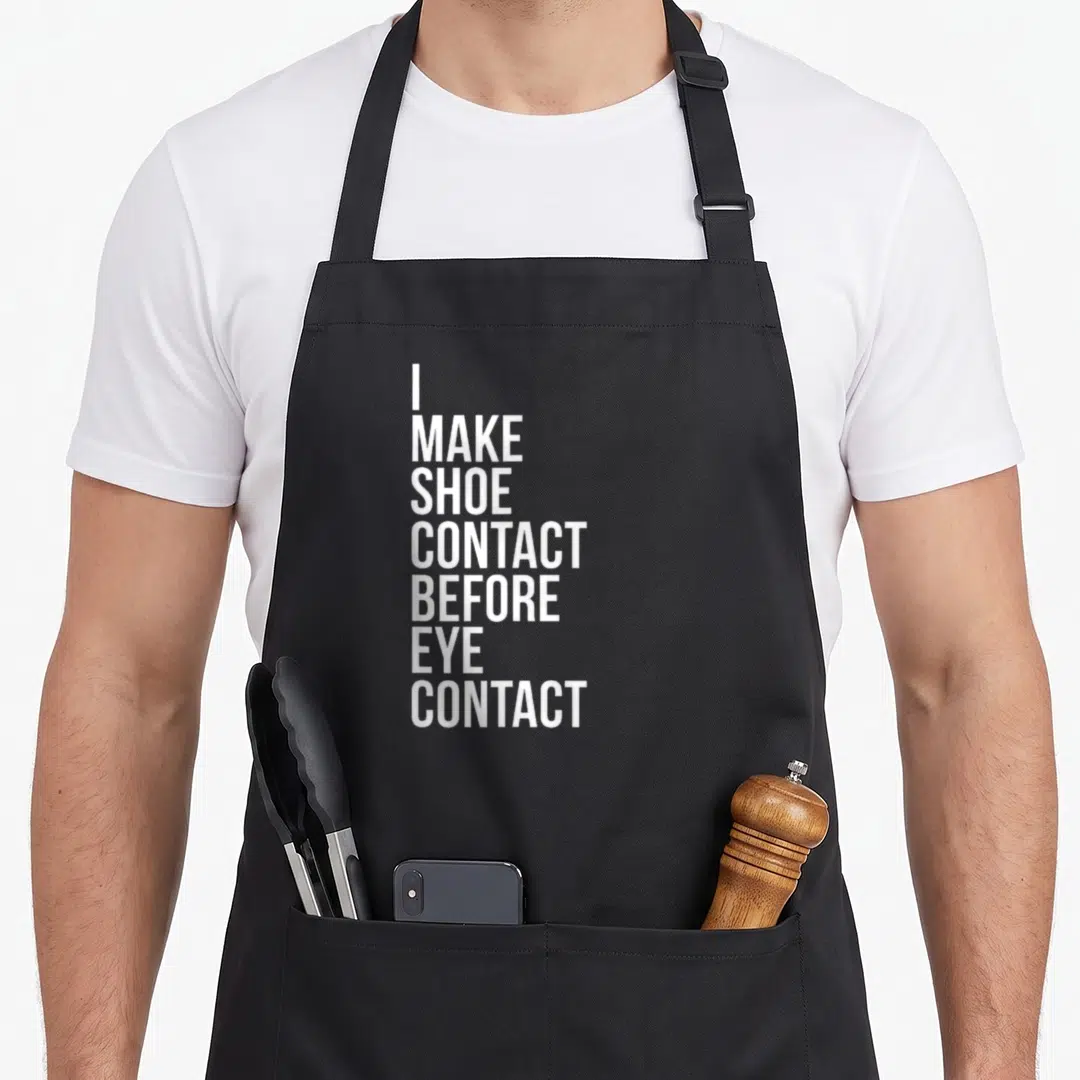 Contact shoe apron - White stacked text reading I MAKE SHOE CONTACT BEFORE EYE CONTACT on grey background — shown on a black cotton canvas apron against a white background