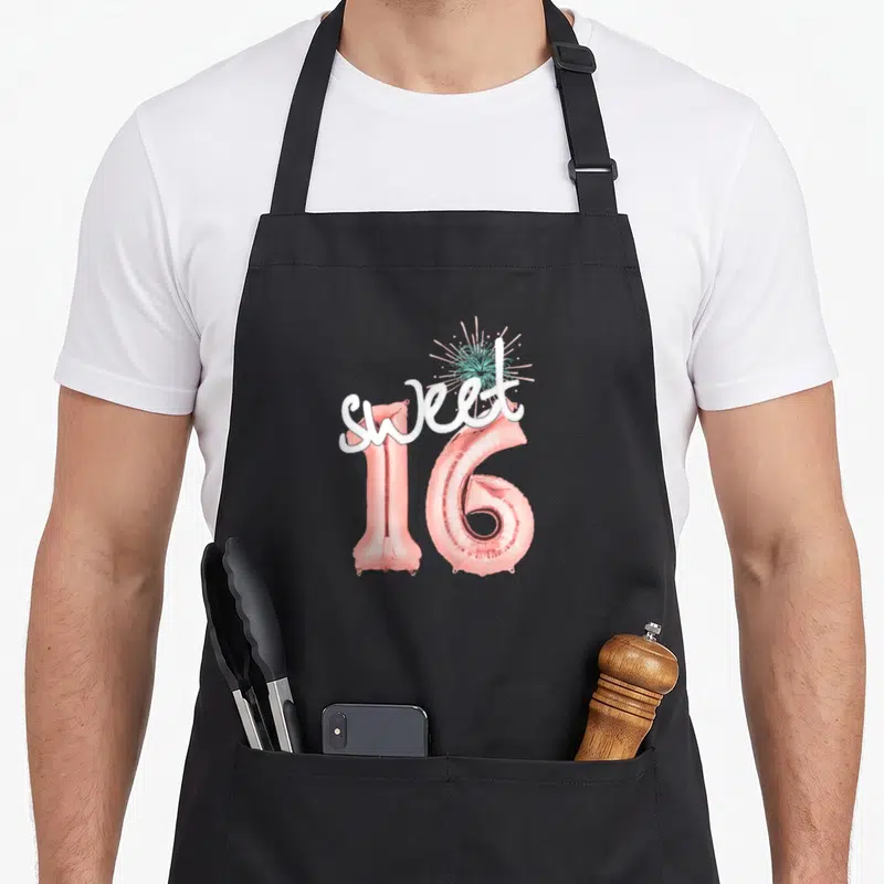 Sweet balloon apron - Rose gold foil balloons forming 16 with white script and teal firework burst — shown on a black cotton canvas apron against a white background