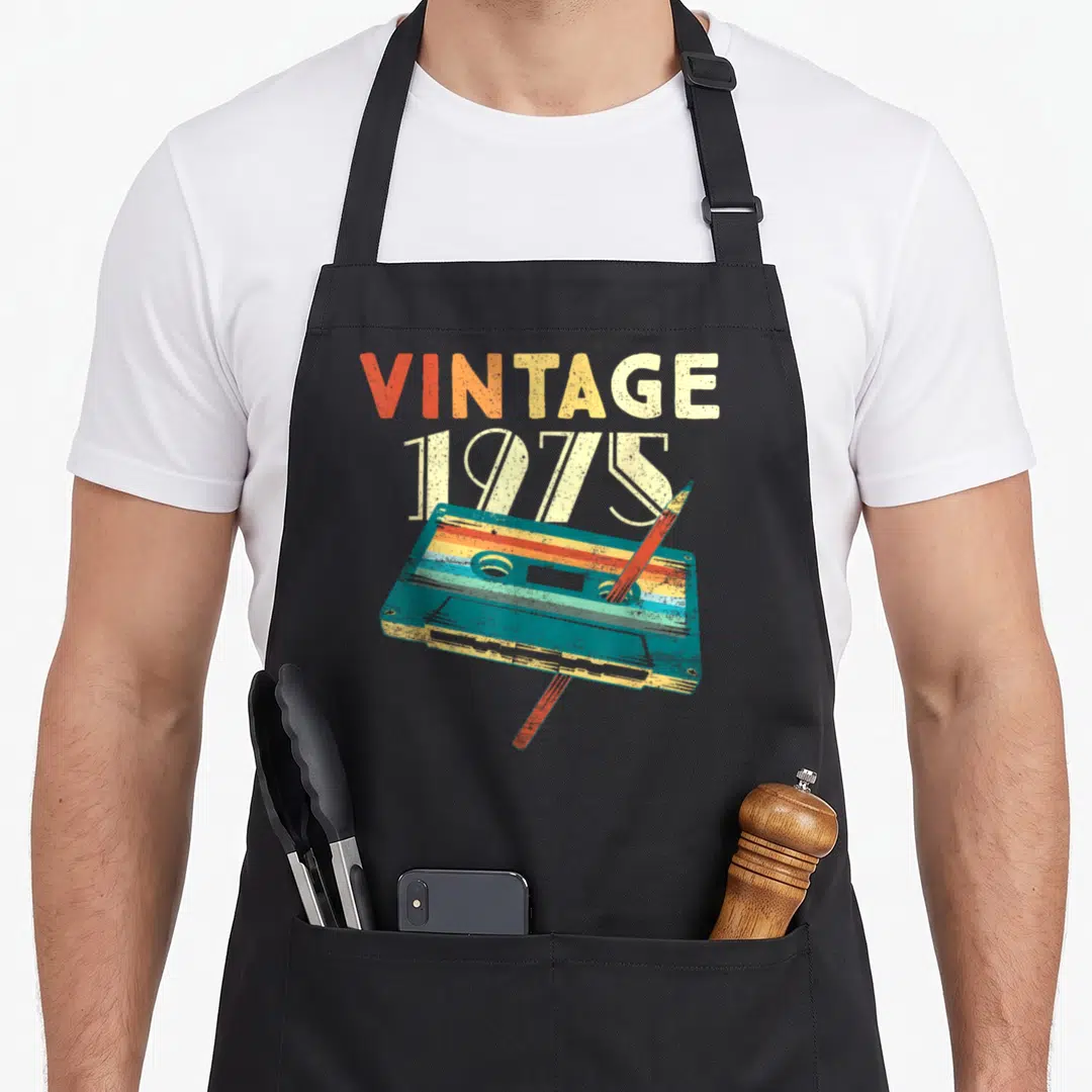 51st birthday apron - Retro cassette tape with VINTAGE 1975 text and diagonal pencil in teal and orange — shown on a black cotton canvas apron against a white background