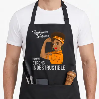 Leukemia chemo warrior apron - Cartoon woman flexing arm with orange headband and text Leukemia Warrior Brave Strong — shown on a black cotton canvas apron against a white background