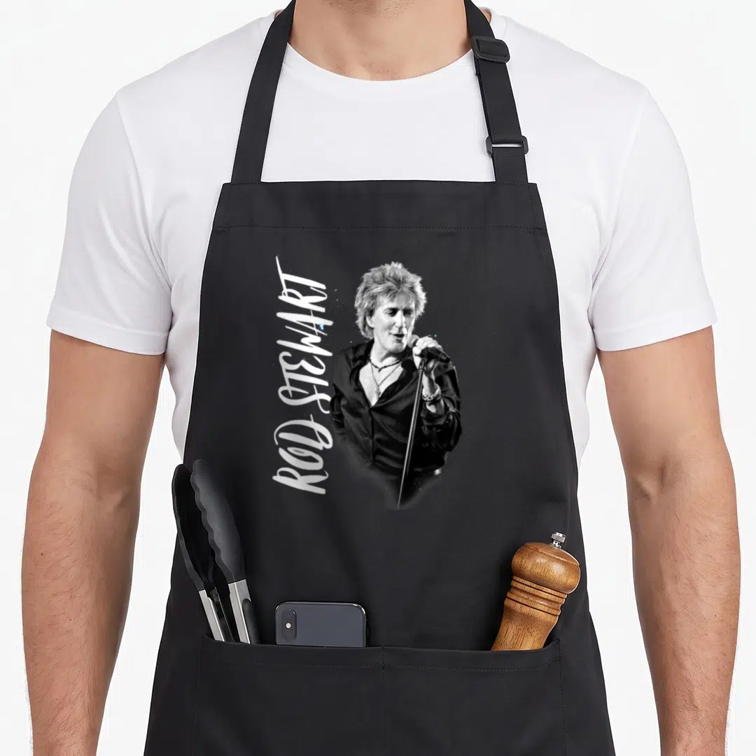 Stewart admat rod apron - Vertical white Rod Stewart text beside black and grey photo of singer holding microphone — shown on a black cotton canvas apron against a white background