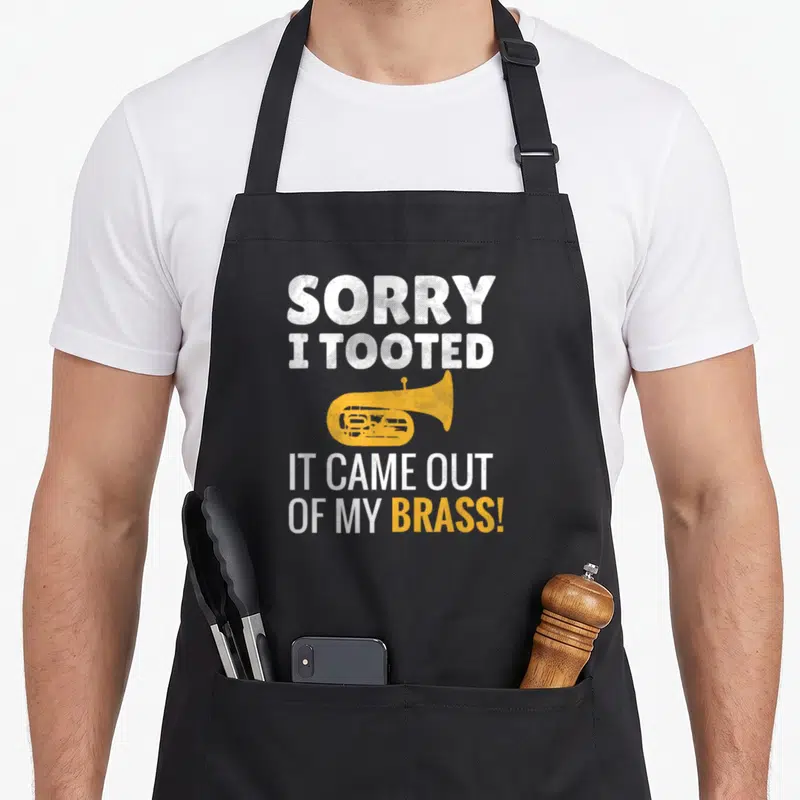 Sorry tuba apron - Yellow tuba graphic between white distressed text reading Sorry I Tooted It Came Out of My Brass — shown on a black cotton canvas apron against a white background