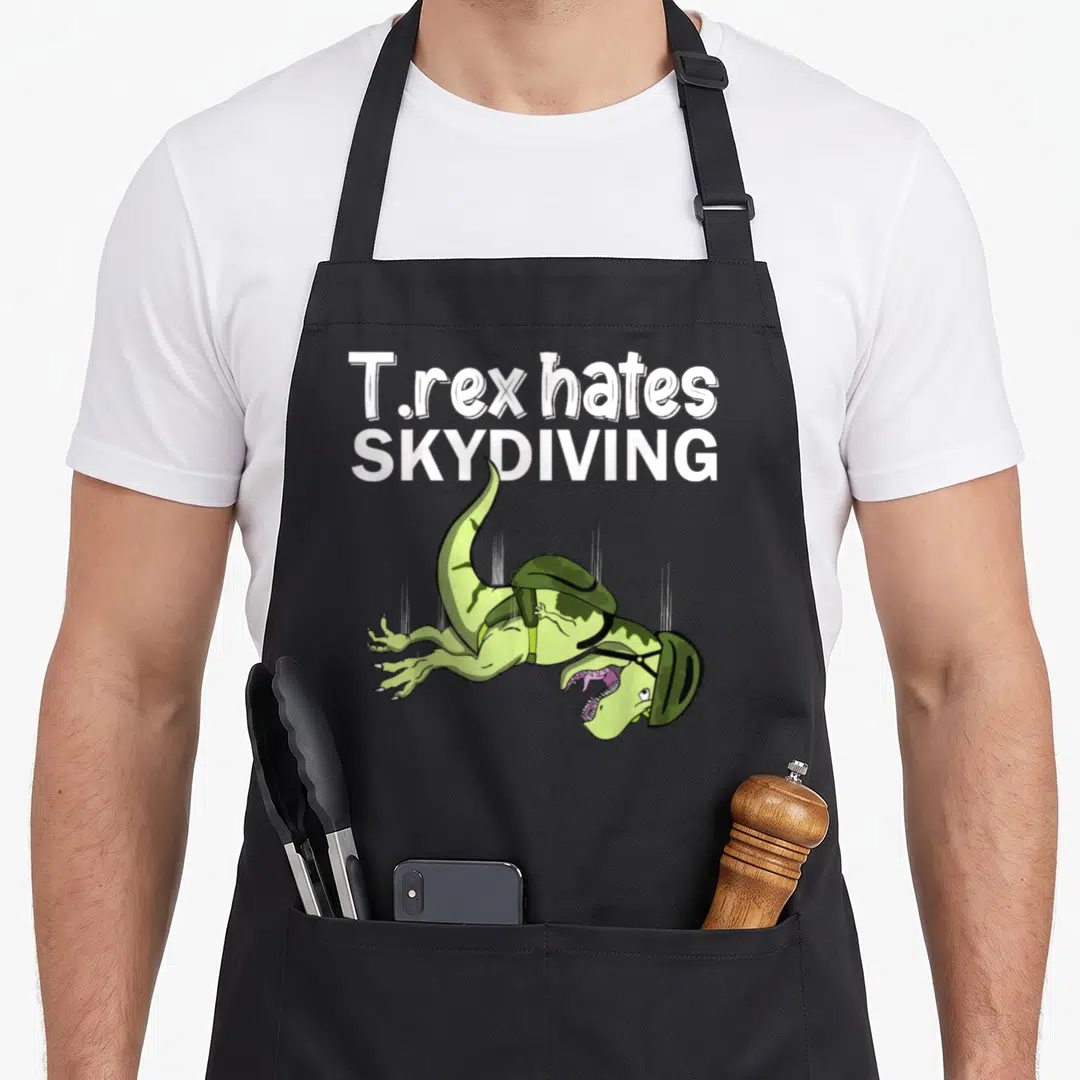 Skydiving t-rex apron - Green cartoon T-rex falling with helmet under white text T.rex hates SKYDIVING — shown on a black cotton canvas apron against a white background