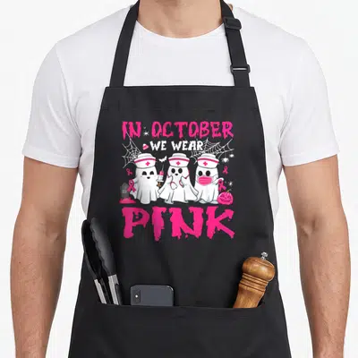 October halloween nurse apron - Three ghost nurses holding pink ribbons and syringes surrounded by spider webs — shown on a black cotton canvas apron against a white background