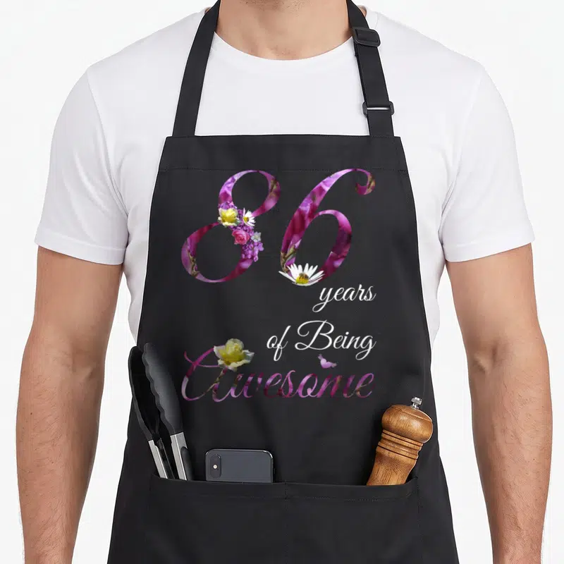 86th floral aprons - Number 86 filled with purple floral pattern and roses, white script text below — shown on a black cotton canvas apron against a white background