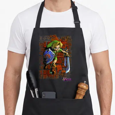 Mask legend apron - Link holding shield and sword with green tunic against distressed orange background and purple logo — shown on a black cotton canvas apron against a white background