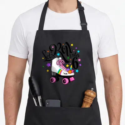 Unicorn roller birthday apron - Let's Roll into 6 text over white roller skate with rainbow stripe and colorful stars — shown on a black cotton canvas apron against a white background