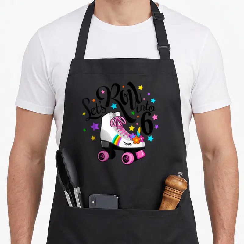 Unicorn roller birthday apron - Let's Roll into 6 text over white roller skate with rainbow stripe and colorful stars — shown on a black cotton canvas apron against a white background
