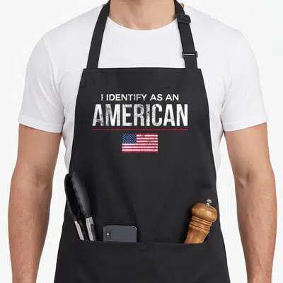 Identify a's aprons - Distressed white text I IDENTIFY AS AN AMERICAN above red line and USA flag — shown on a black cotton canvas apron against a white background