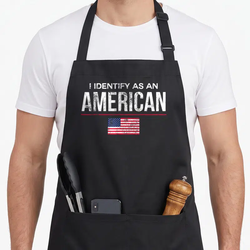 Identify a's aprons - Distressed white text I IDENTIFY AS AN AMERICAN above red line and USA flag — shown on a black cotton canvas apron against a white background