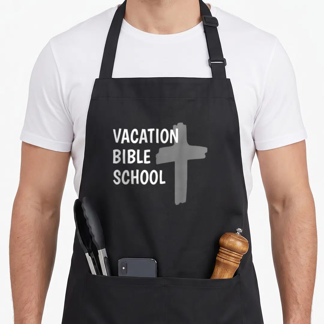 Vacation church aprons - White stacked VACATION BIBLE SCHOOL text beside a faded grey cross on dark fabric — shown on a black cotton canvas apron against a white background