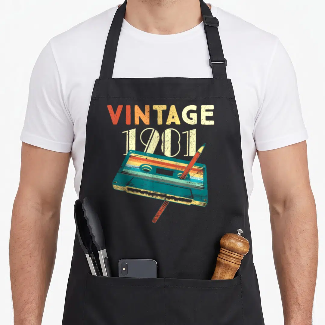 Cassette 45th birthday apron - Retro cassette tape and pencil below stacked VINTAGE 1981 text in teal and orange — shown on a black cotton canvas apron against a white background