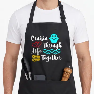 Cruise newlywed aprons - Stacked white text Cruisin Through Life Together with cyan ship red hearts waves yellow flip — shown on a black cotton canvas apron against a white background