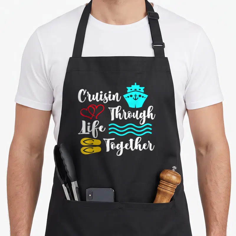 Cruise newlywed aprons - Stacked white text Cruisin Through Life Together with cyan ship red hearts waves yellow flip — shown on a black cotton canvas apron against a white background