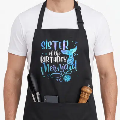 Sister birthday mermaid squad family aprons - Stacked text SISTER of the BIRTHDAY Mermaid with blue purple scaled tail — shown on a black cotton canvas apron against a white background