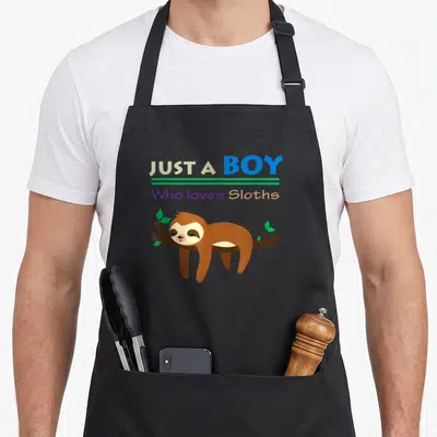 Funny lazy sloth apron - Text Just A Boy Who loves Sloths above cartoon sloth lying on branch — shown on a black cotton canvas apron against a white background