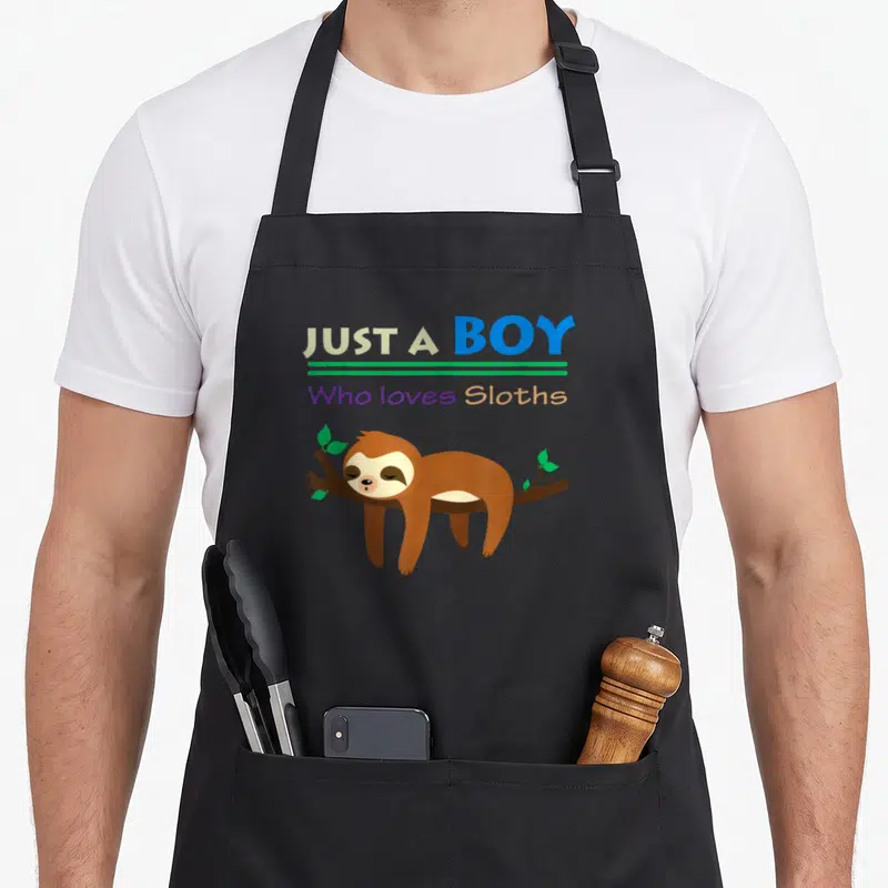 Funny lazy sloth apron - Text Just A Boy Who loves Sloths above cartoon sloth lying on branch — shown on a black cotton canvas apron against a white background