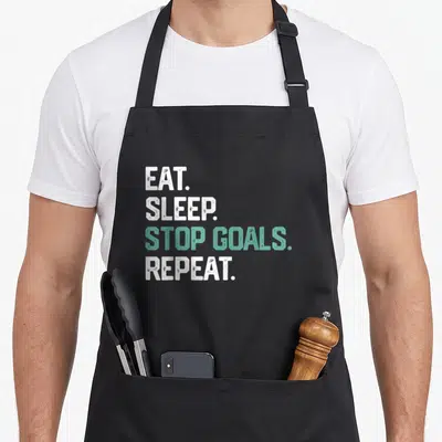 Eat soccer boys apron - Stacked white and teal text reading EAT SLEEP STOP GOALS REPEAT on black fabric — shown on a black cotton canvas apron against a white background