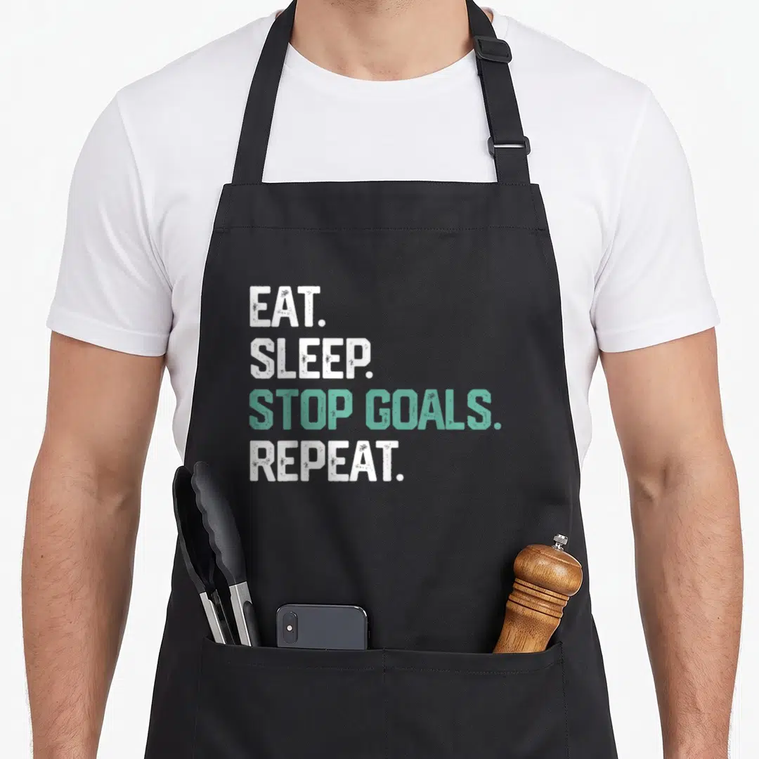 Eat soccer boys apron - Stacked white and teal text reading EAT SLEEP STOP GOALS REPEAT on black fabric — shown on a black cotton canvas apron against a white background