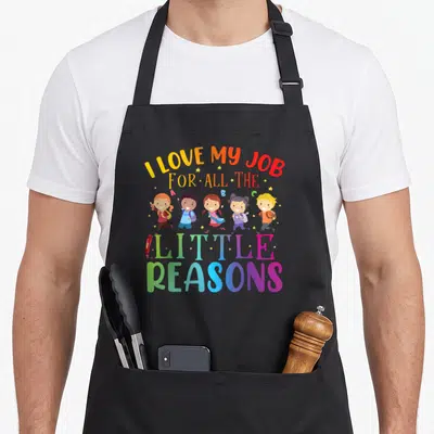 Reason teaching apron - Five cartoon children running under I LOVE MY JOB FOR ALL THE LITTLE REASONS text with pencil — shown on a black cotton canvas apron against a white background